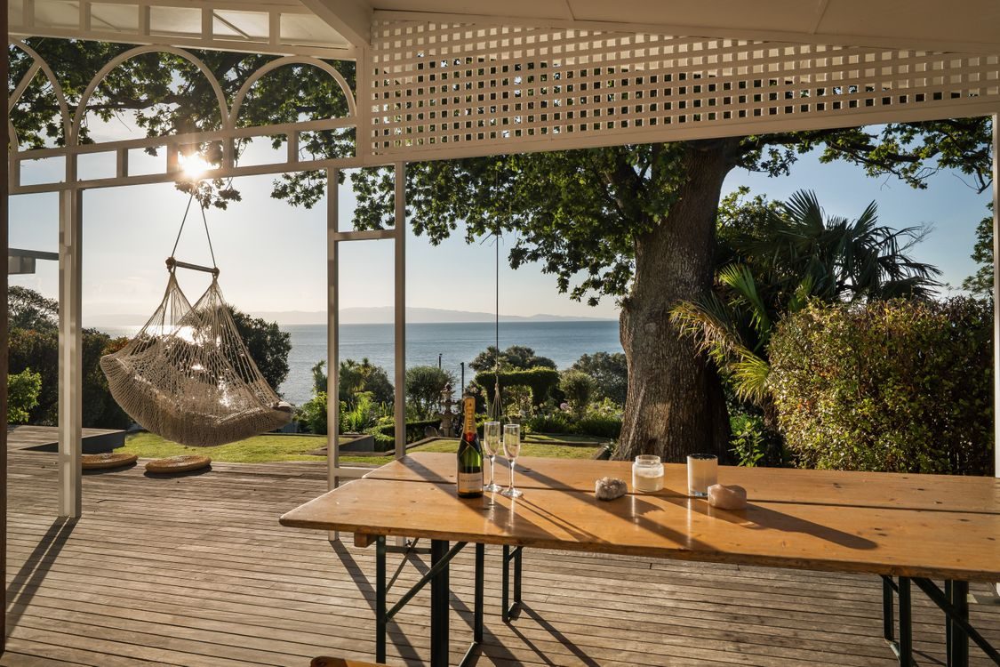 The covered deck and outdoor table beneath a large tree, sea beyond